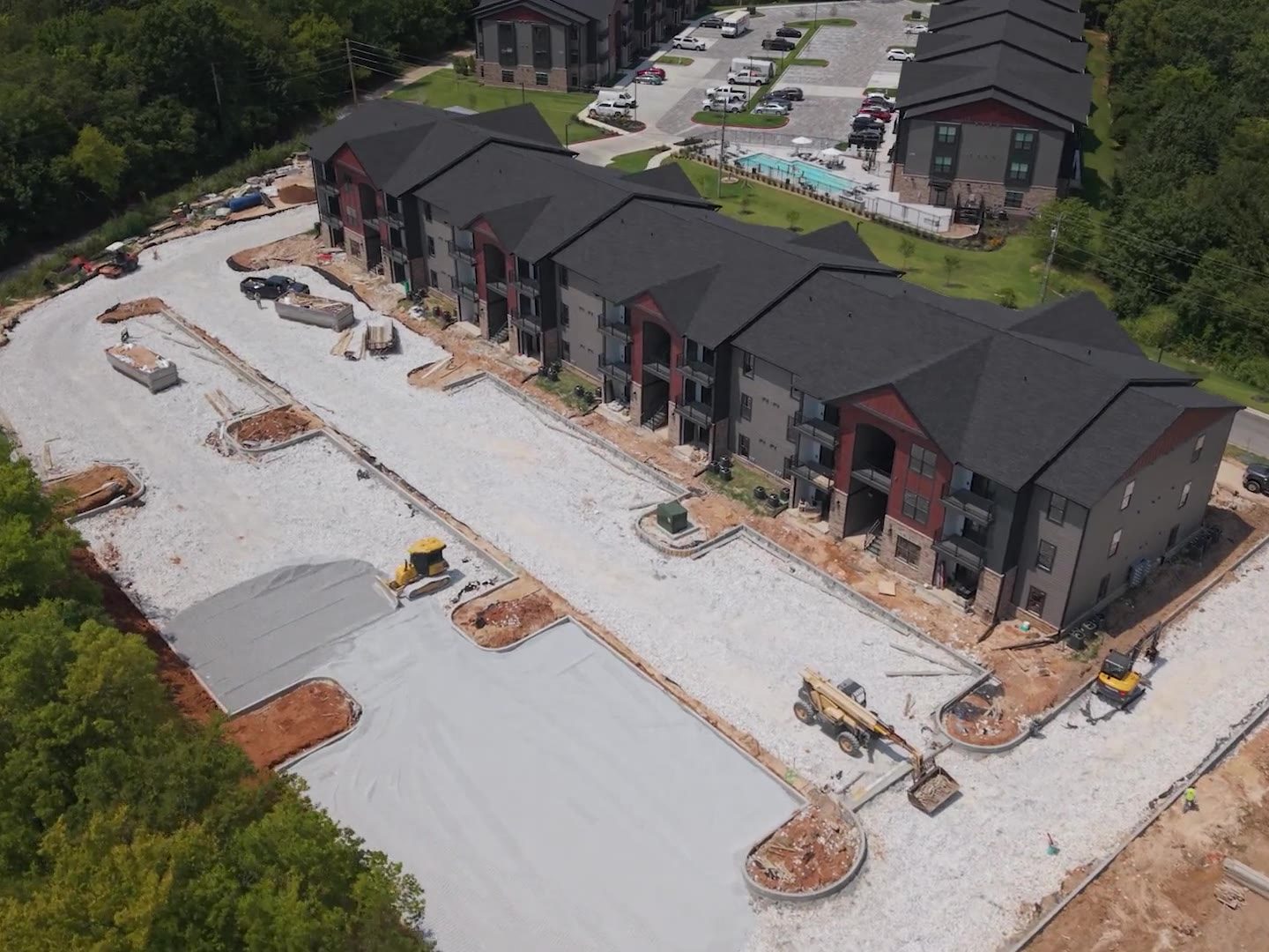 Aerial view of a multi-family apartment complex under construction, showing freshly poured concrete, gravel sub-base and grading equipment alongside completed buildings