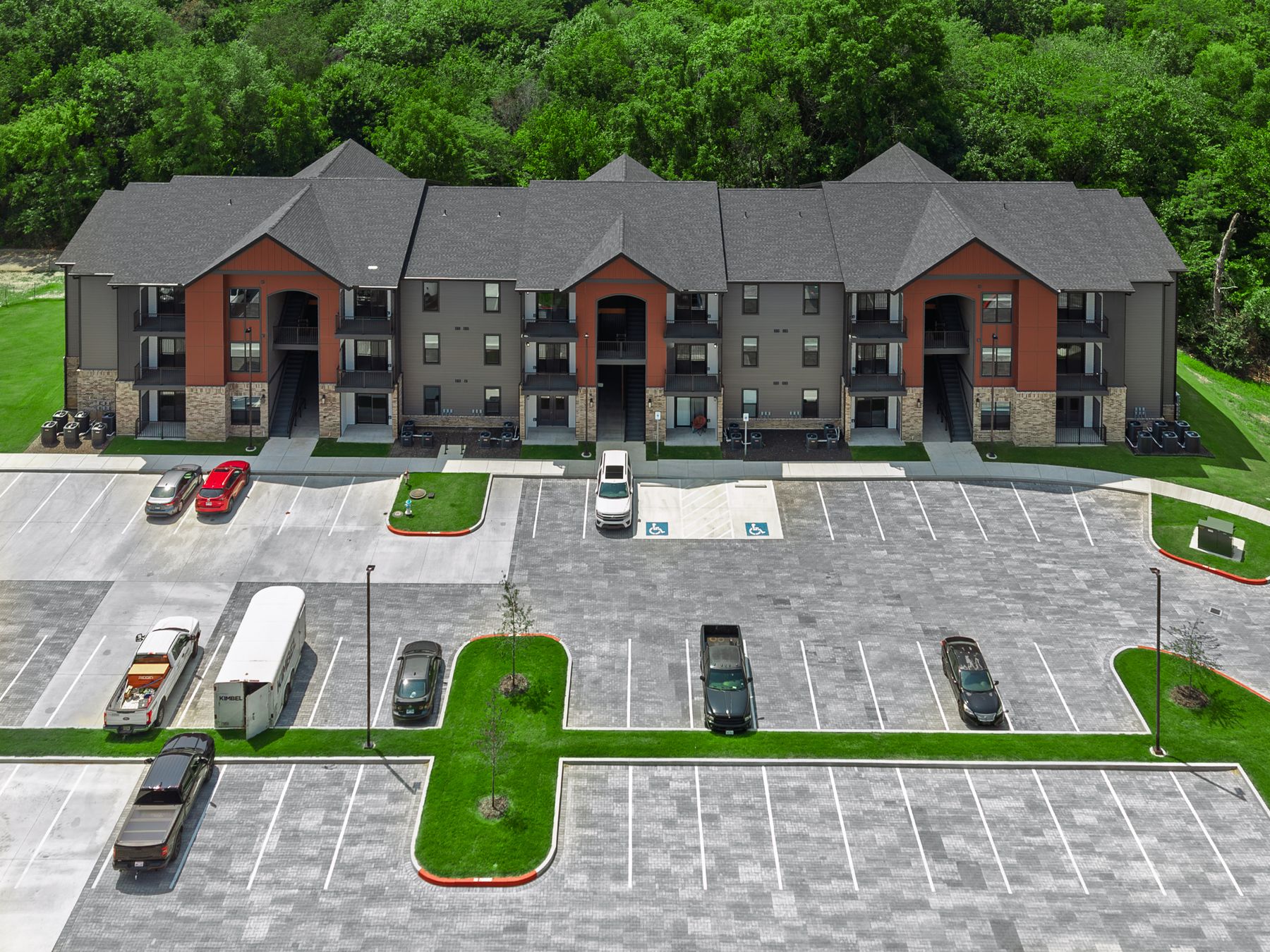 Elevated aerial of a three-story apartment building with rust-red gables and stacked balconies, behind its paver parking lot