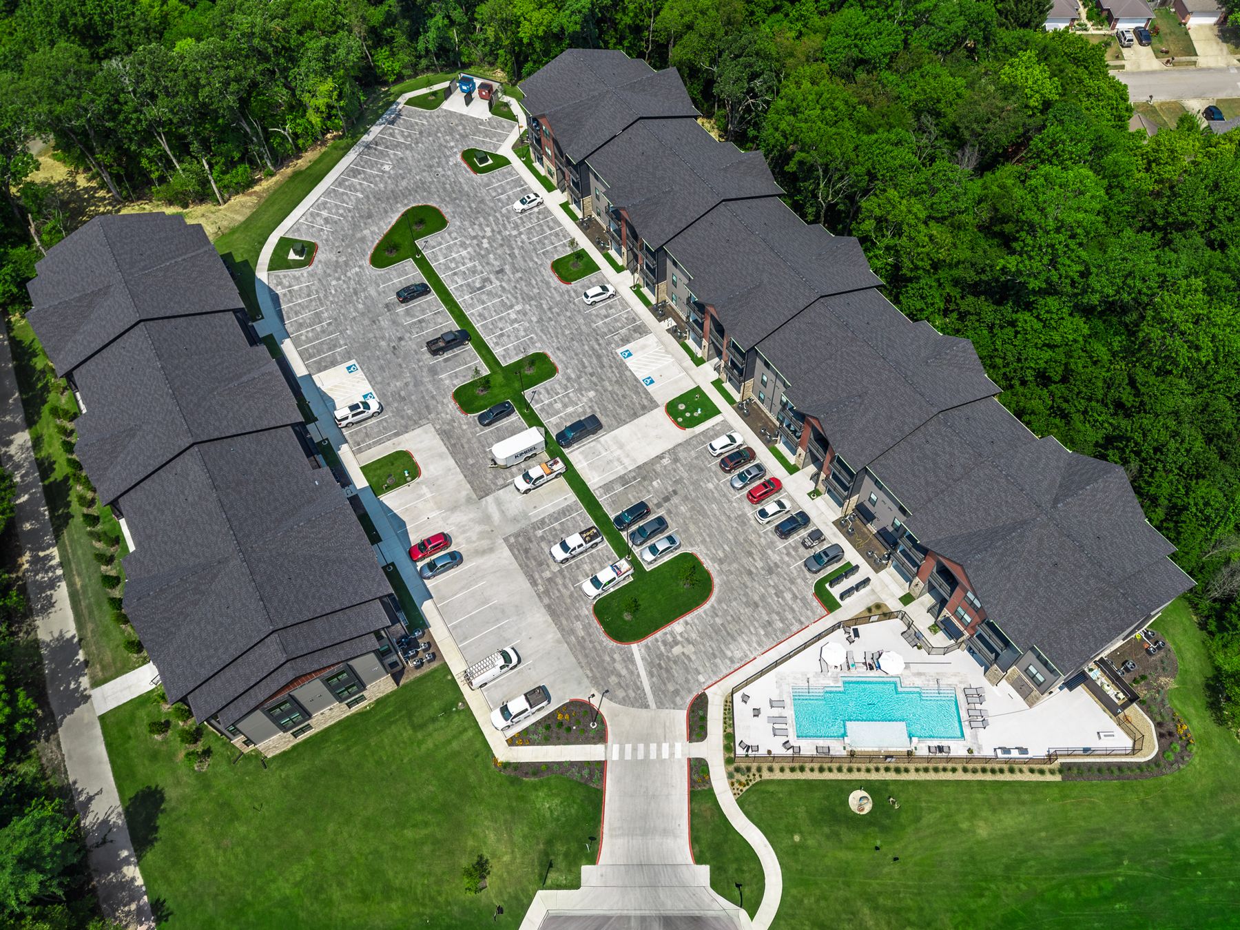 Overhead aerial of a new apartment complex: two three-story buildings either side of a paver parking lot, with a pool at the entrance and woods all around
